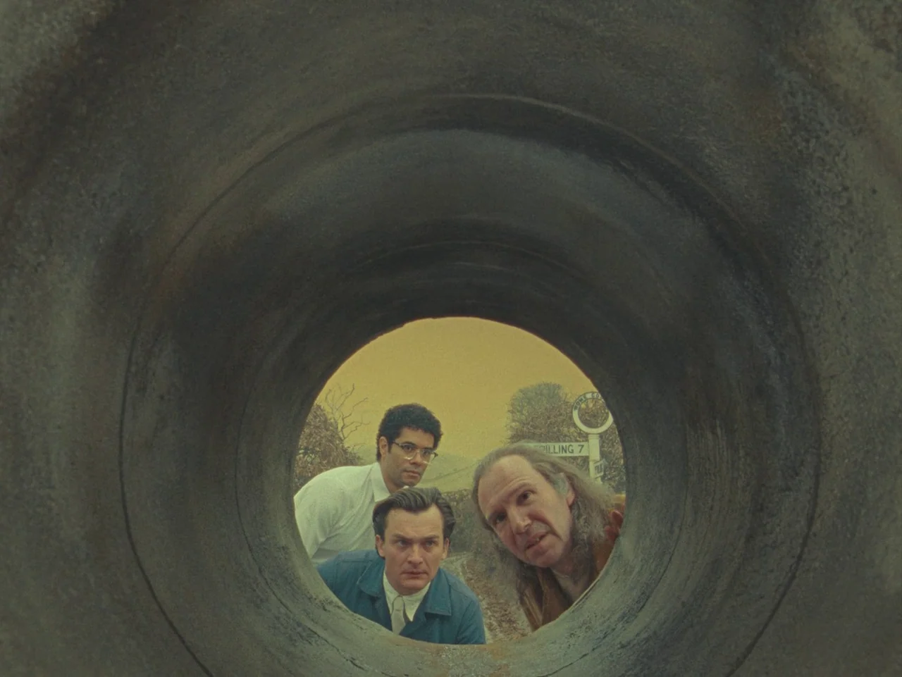 The Rat Catcher (2023) movie still: medium — Three men peer through the opening of a massive concrete drainage pipe outdoors. A directional sign…