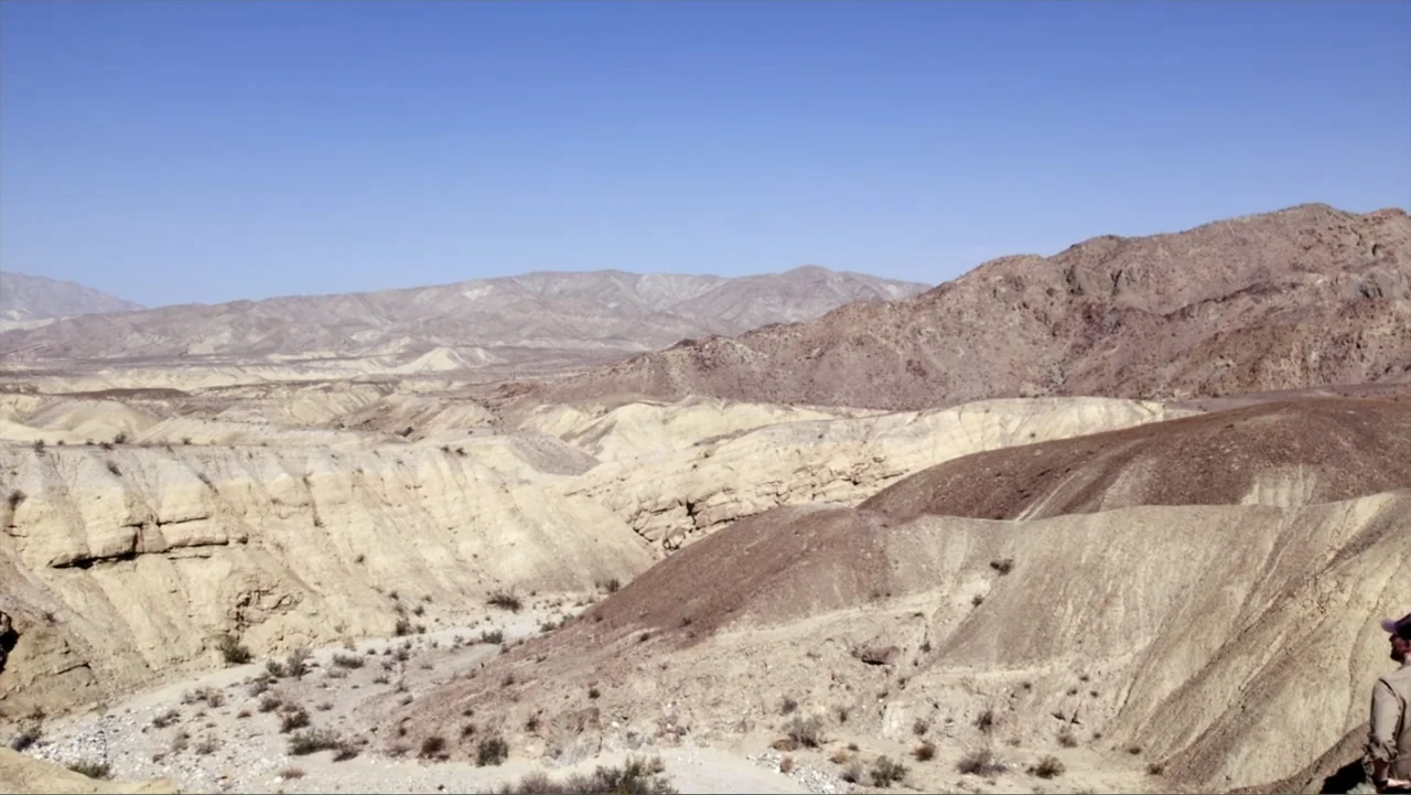 The Atlantis Puzzle (2024) movie still: wide — An arid, rocky desert landscape stretches out with jagged beige and brown geological formations. A…