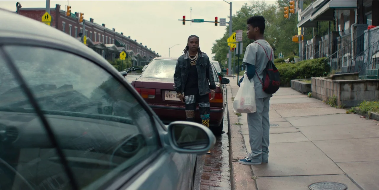 Charm City Kings (2020) movie still: wide — An urban residential street lined with row houses under an overcast sky. Two young men converse on the…