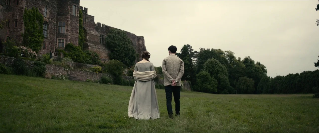 The King movie still: fullbody — A stone castle with overgrown ivy overlooks a sloping green meadow. A young woman and man walk away from the camera…