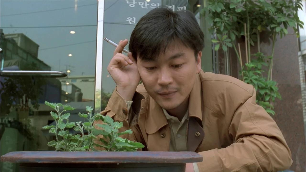 The Day a Pig Fell Into the Well (1996) movie still: medium — A man sits outdoors in front of a building with glass doors and signage. He leans…
