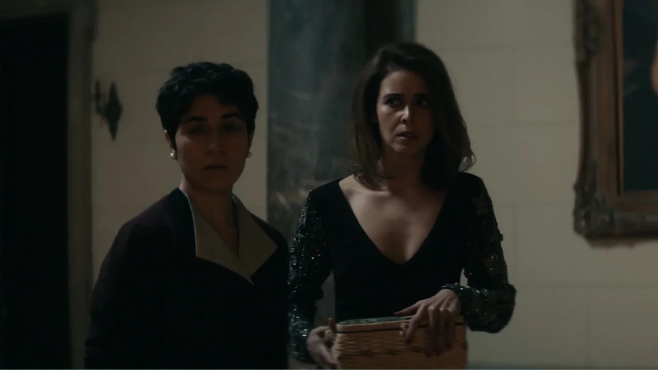 Bel Canto (2018) movie still: medium — A dimly lit indoor hallway features pale walls and a framed painting. Two women in formal attire walk side by…