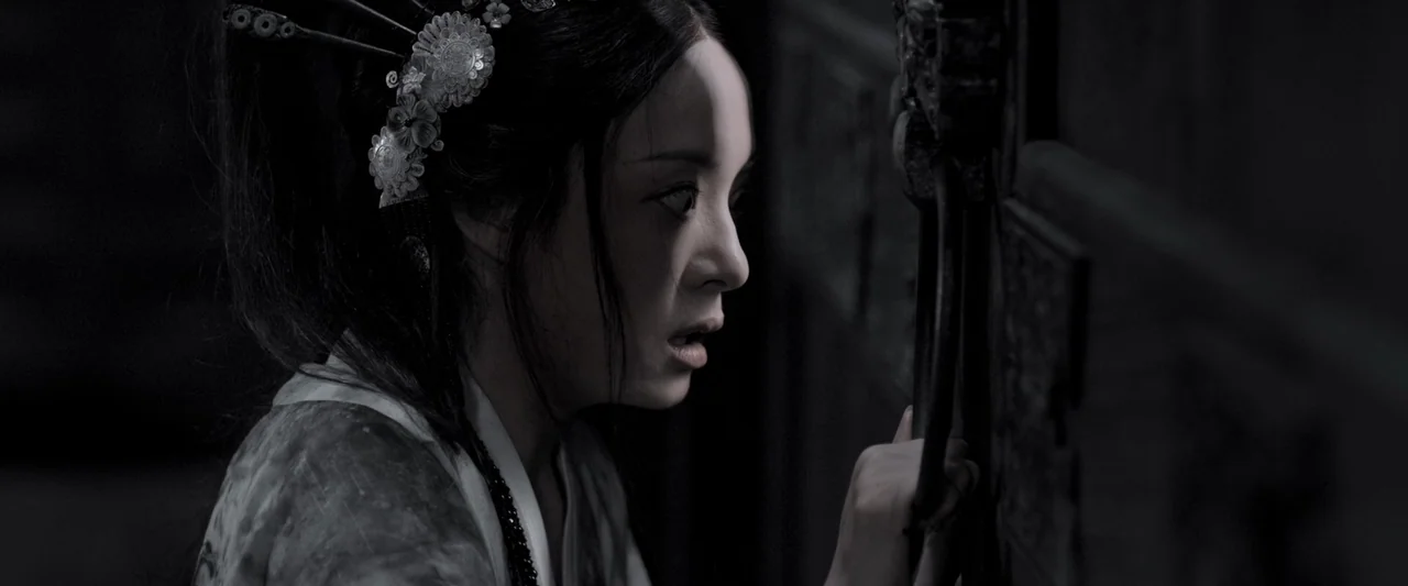 Shadow (2018) movie still: closeup — A woman in traditional attire stands near a dark wooden structure with ornate carvings. She stares through a…