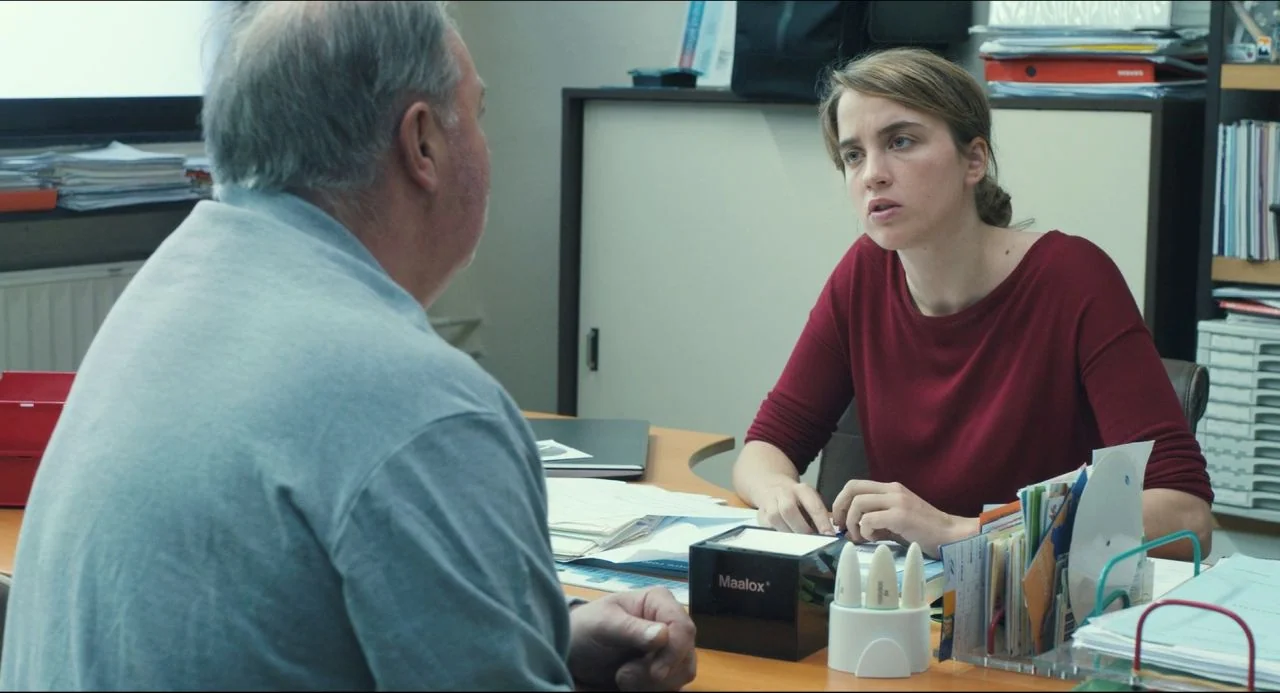 The Unknown Girl (2016) movie still: medium — A cluttered professional work space with shelves filled with files and a desk surface. A young woman…