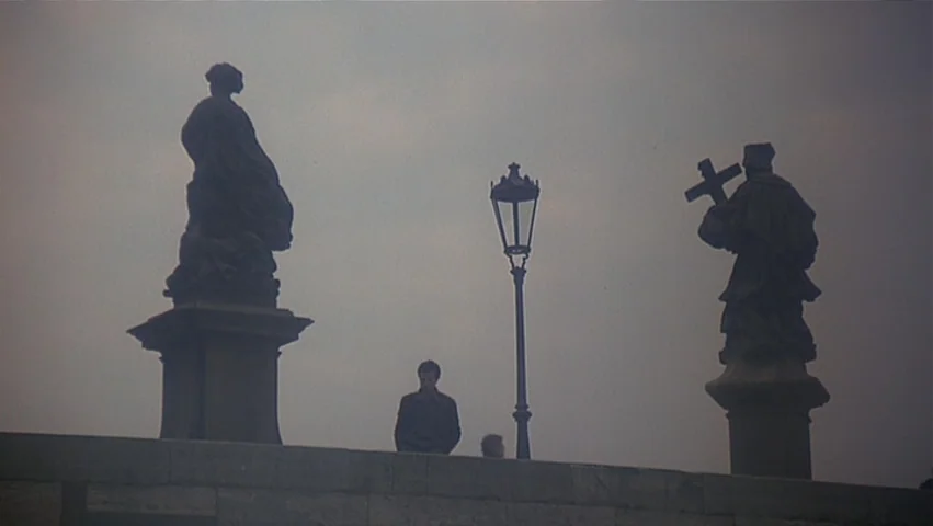 The Unbearable Lightness of Being (1988) movie still: wide — A stone bridge features two large religious statues flanking a central iron lamp post.…