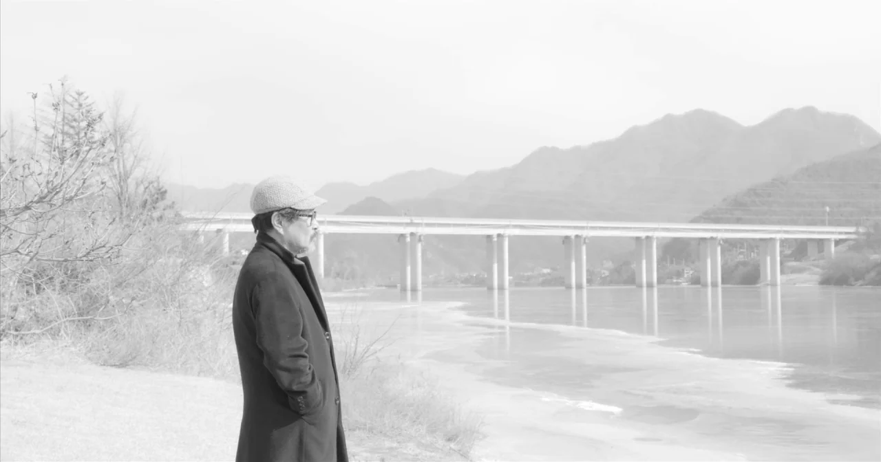 Hotel By The River movie still: fullbody — A man stands on the bank of a partially frozen river during daytime. A concrete bridge spans the distance…