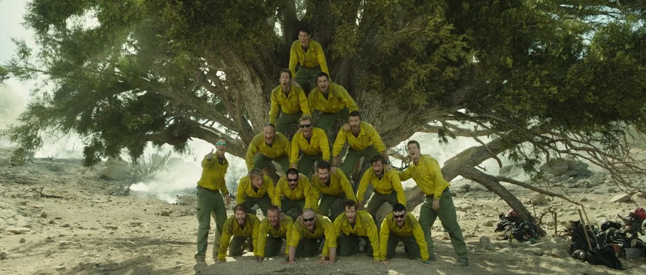 Only The Brave (2017) movie still: wide — A group of wildfire firefighters in uniform poses in a human pyramid formation outdoors. They are arranged…