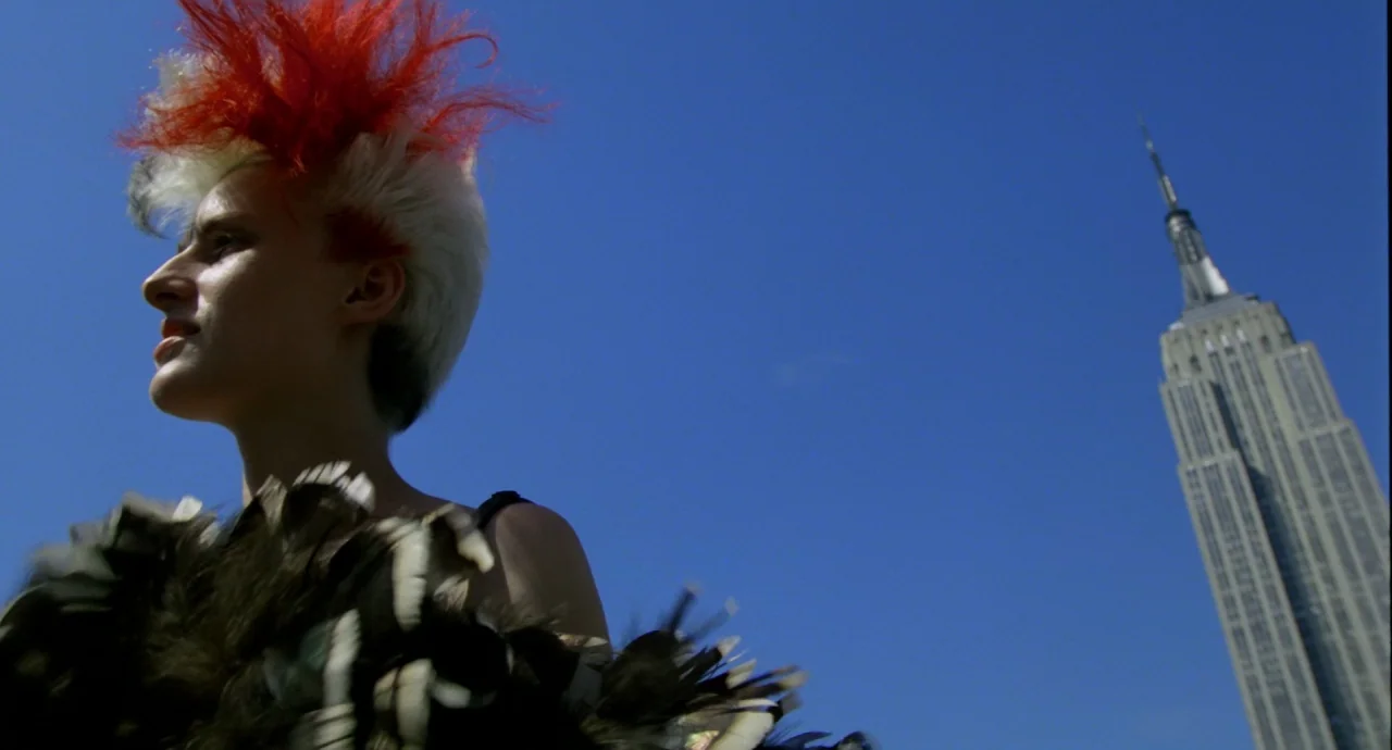 Liquid Sky (1982) movie still: medium — A person with a punk hairstyle stands against a deep blue sky backdrop. The Empire State Building is visible…