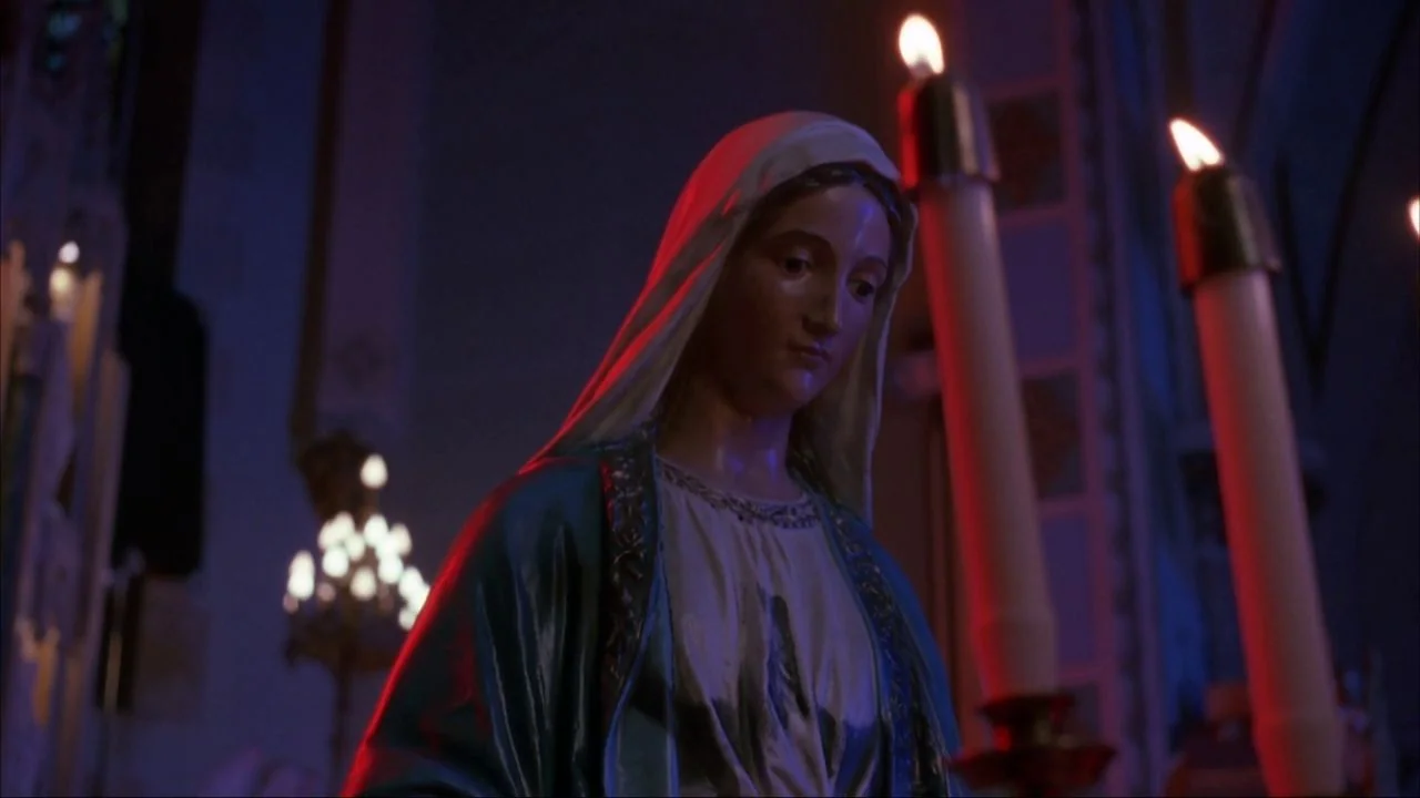 Bad Lieutenant (1992) movie still: medium — A religious statue stands inside a dimly lit space illuminated by tall candles. The figure wears blue…