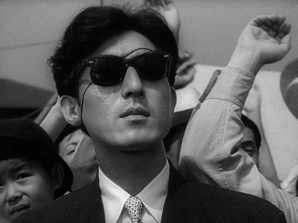 Godzilla (1954) (1954) movie still: closeup — A man stands in the middle of a dense crowd during the day. He wears dark sunglasses and looks upward…