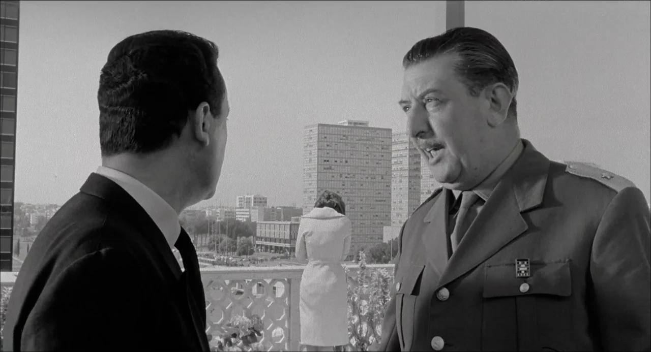 Il Boom (1963) movie still: medium — A stone terrace overlooks a city with tall modernist buildings in the background. Two men engage in…