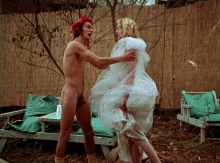 Desperate Living (1977) movie still: fullbody — A backyard space features a reed fence and outdoor wooden furniture with green cushions. A nude man…