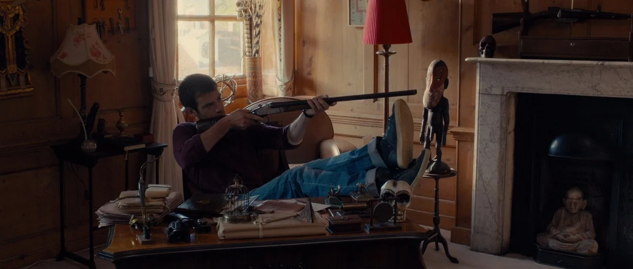 Road Games (2015) movie still: medium — A study features wood-paneled walls and a desk cluttered with antiques and small objects. A man with short…