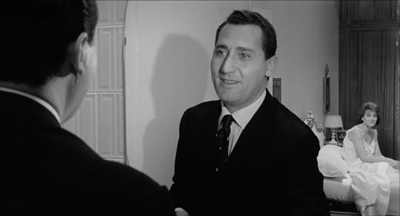 Il Boom (1963) movie still: medium — A well-lit room contains a seated woman and two standing men in suits. One man speaks while positioned in the…