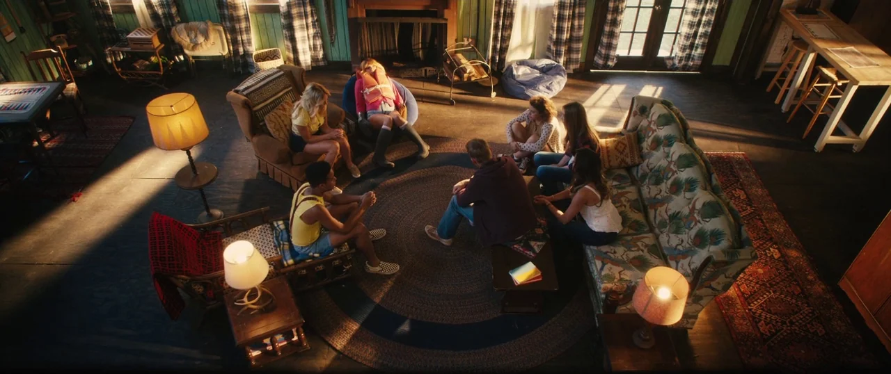 Final Girls (2015) movie still: wide — A spacious rustic living room with wood paneled walls contains a central circular rug. Seven young adults sit…