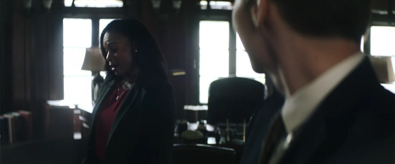 Truth or Dare (2018) movie still: medium — A dark office features a large desk and window with bright light spilling through. A woman in a maroon…