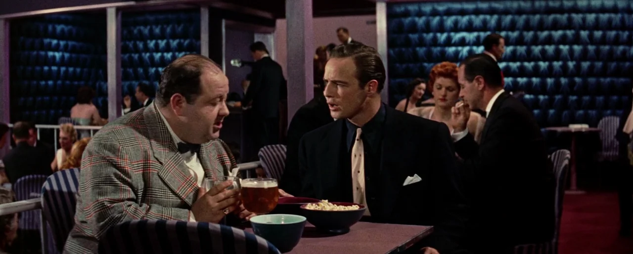 Guys and Dolls (1955) movie still: medium — A dimly lit nightclub features blue padded walls and patrons seated at small tables. Two men in suits…