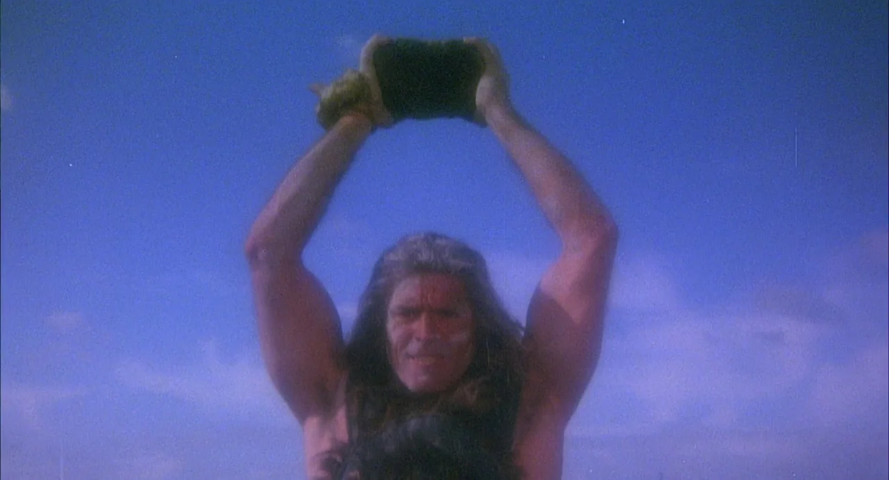 Conquest (1983) movie still: medium — A muscular man stands in an outdoor environment against a clear, expansive blue sky. He holds a large,…