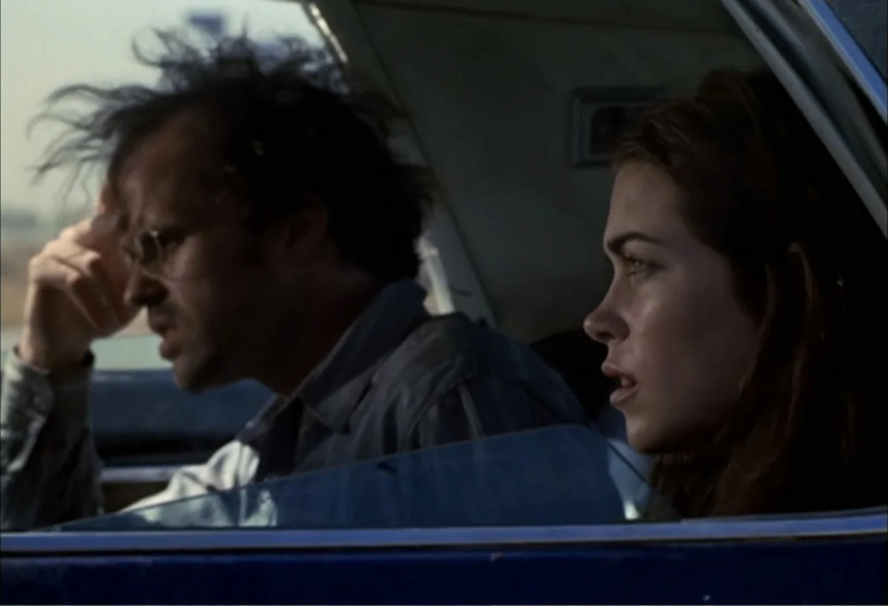 Black Cat Run (1998) movie still: medium — Interior of a moving vehicle during daylight hours with bright exterior reflections visible. A man drives…