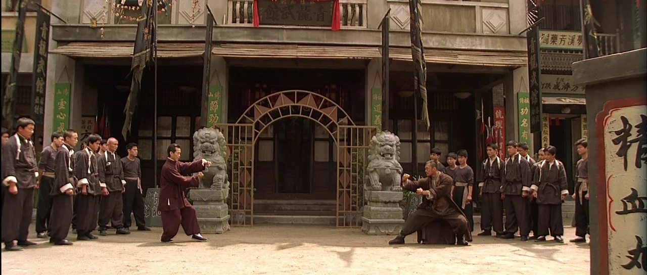 Ip Man (2008) movie still: wide — A dusty outdoor courtyard sits before a traditional stone building with ornate Chinese architecture. Two men in…