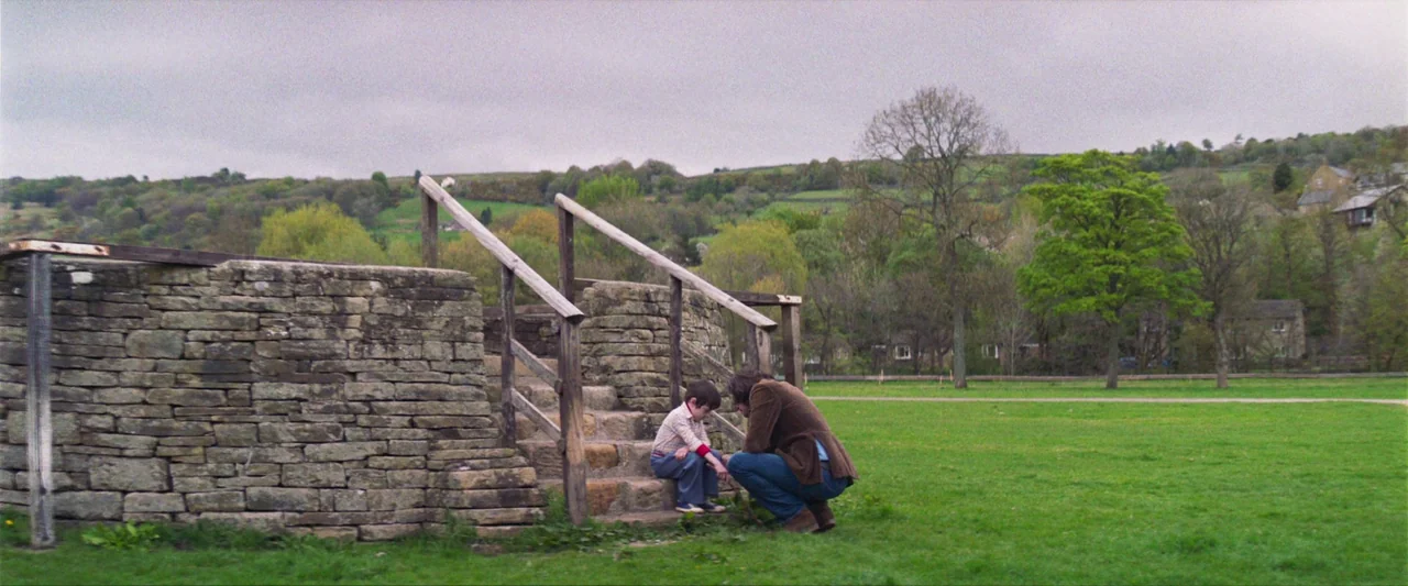 Starve Acre (2023) movie still: wide — A man and a young boy interact near stone steps in a rural field. The background features rolling green hills…