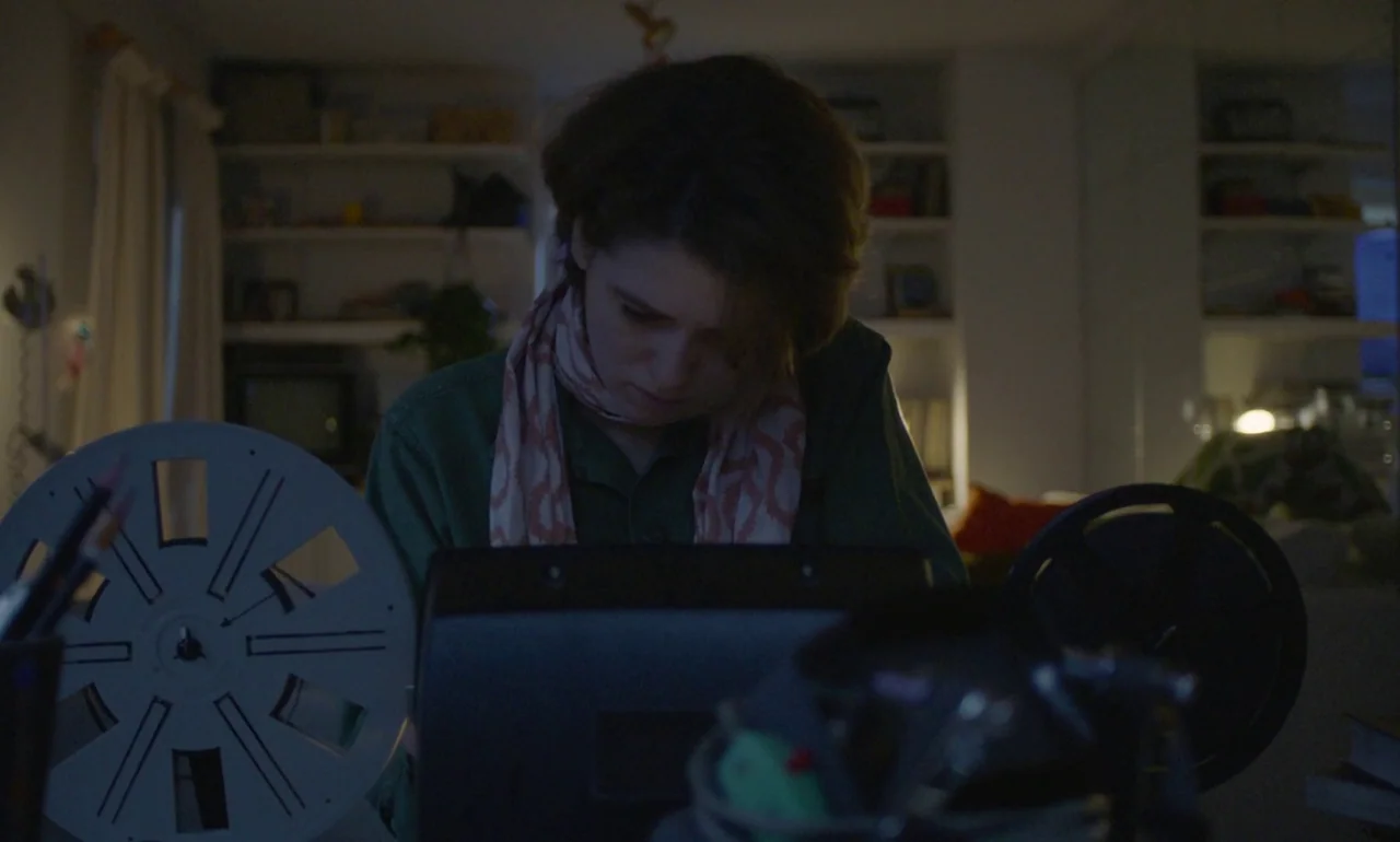 The Souvenir (2019) movie still: medium — A woman in a dark shirt works at a desk containing vintage film editing equipment. Behind her are…
