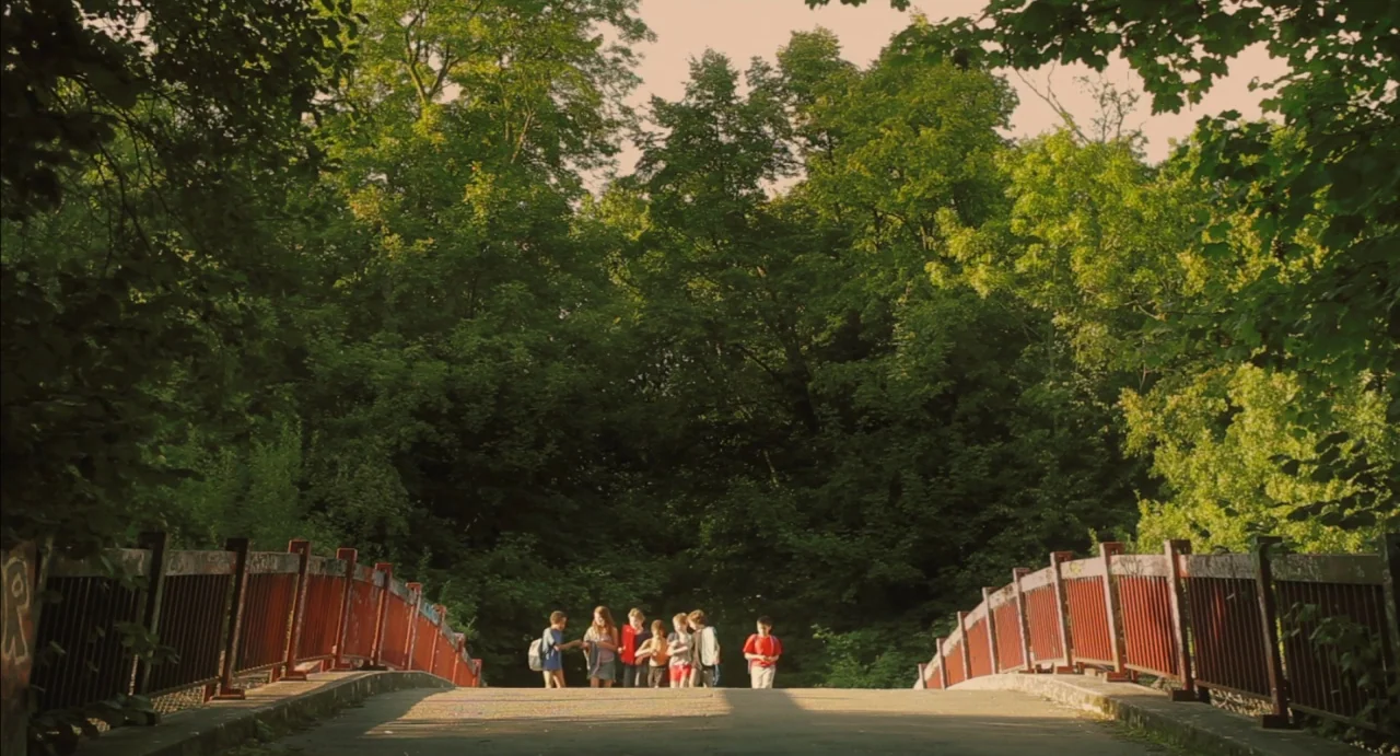 Tomboy (2011) movie still: wide — A group of six children walks across a weathered pedestrian bridge in a park. Dense green forest foliage fills the…
