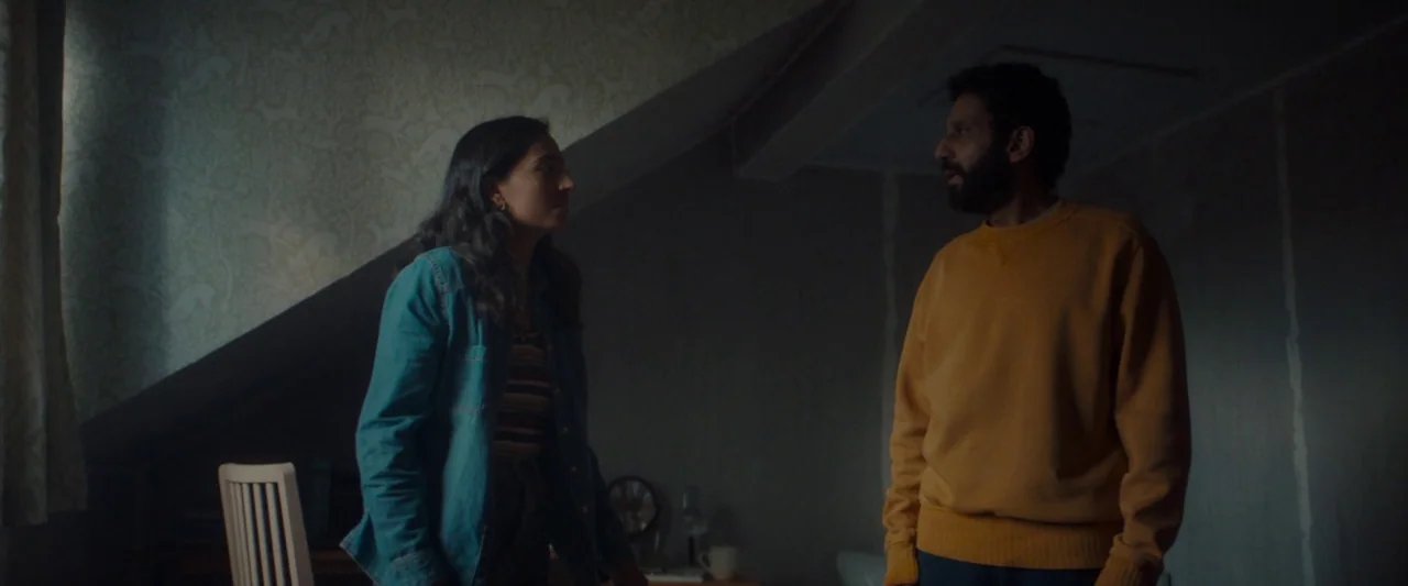 Ali & Ava (2021) movie still: medium — A dark, dimly lit attic space with slanted walls covered in patterned wallpaper. A man in a yellow sweatshirt…