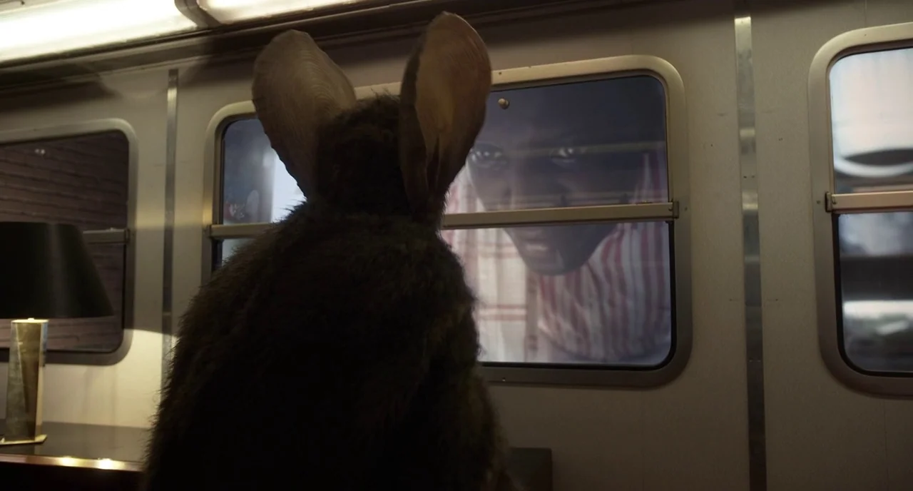 Mood Indigo (2013) movie still: medium — A dark, furred rabbit creature faces a window inside a metal-walled passenger car. The glass reflects a man…