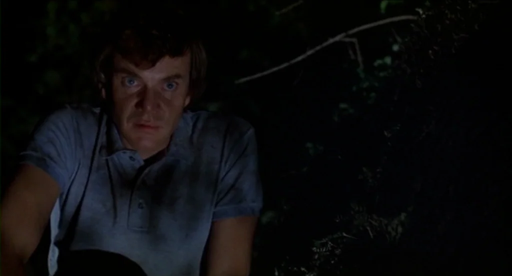 Cat People (1982) movie still: medium — A dark, wooded environment at night features dense foliage in the background. A man crouches while looking…