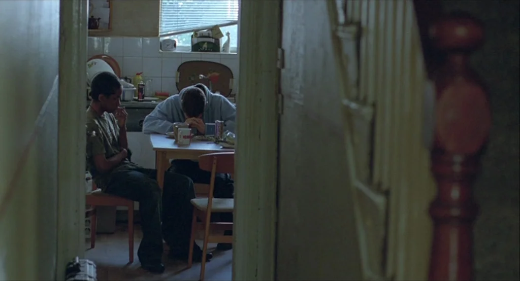 Looking for Eric (2009) movie still: medium — A cluttered residential kitchen with tiled walls and a small wooden table. Two young men sit together,…