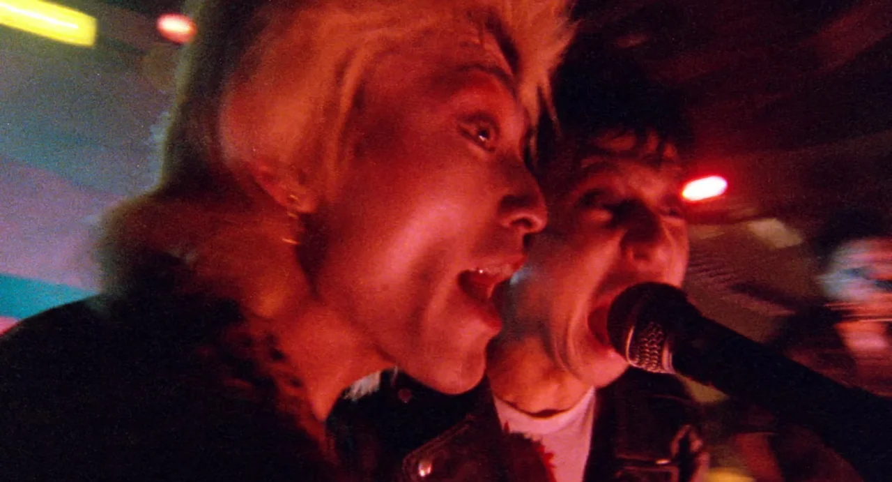 Burst City (1982) movie still: medium — A dimly lit nightclub performance space bathed in intense red light and shadows. Two musicians stand closely…