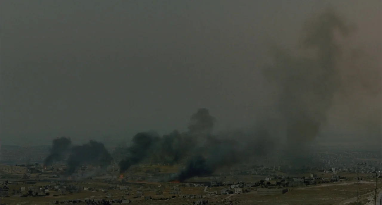 Incendies movie still: establishing — A wide, arid landscape with a scattered town under a heavy, overcast grey sky. Several columns of dark smoke…