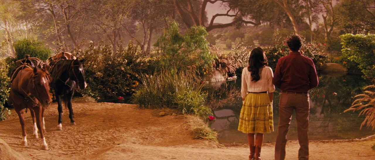Casa De Mi Padre (2012) movie still: wide — A rural landscape features a calm pond surrounded by dense trees and wildflowers. A man and woman stand…