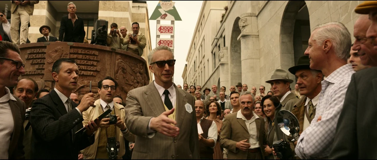 Ferrari (2023) movie still: medium — An outdoor public square features a crowd of men in 1950s business attire. The central man reaches out his hand…
