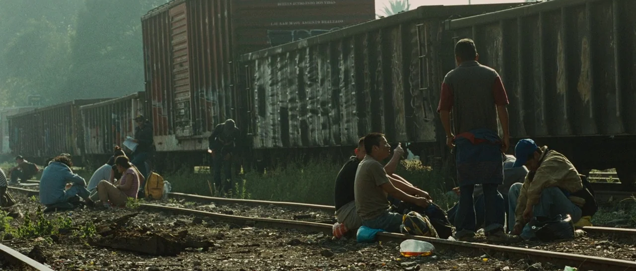 Sin Nombre (2009) movie still: wide — A group of people sits among gravel and weeds beside parked rusted metal freight cars. The subjects are…
