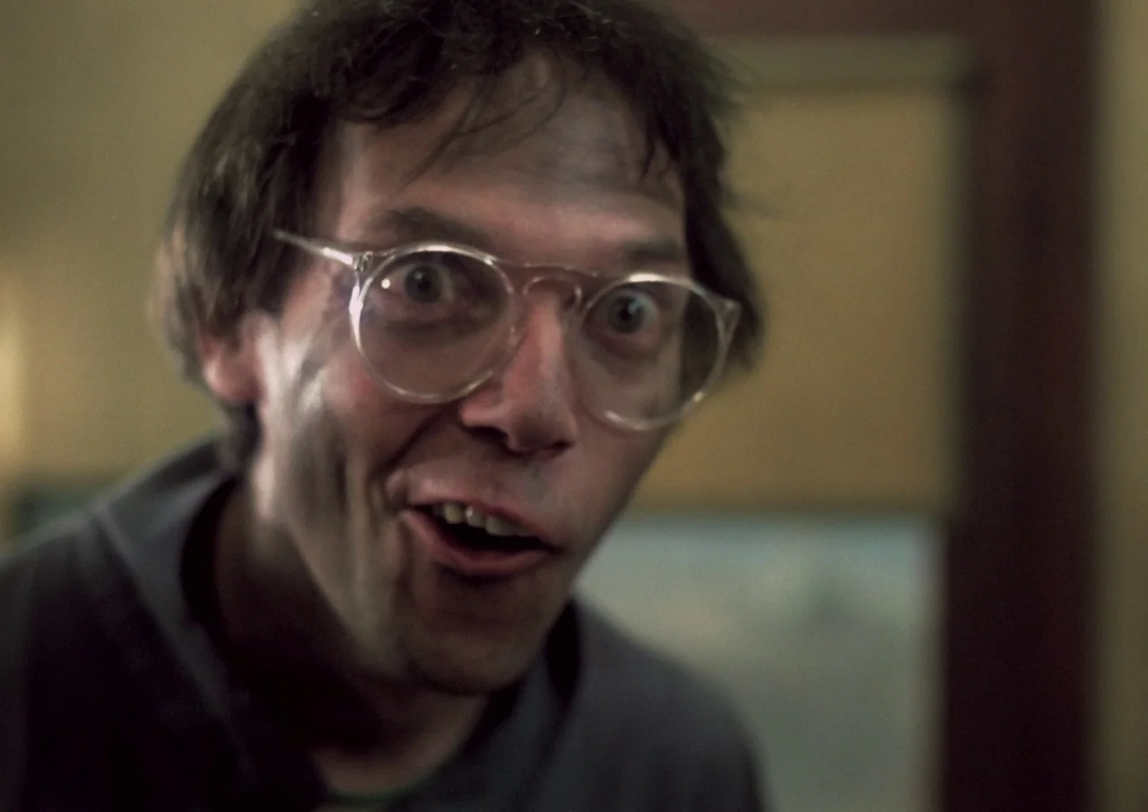 Human Highway (1982) movie still: closeup — The frame shows a close view of a man's face in a dim room. He looks directly at the camera with a…