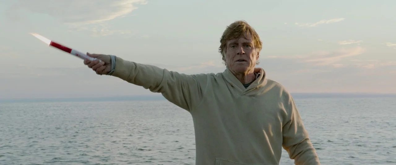 All Is Lost (2013) movie still: medium — An older man stands on open water under a pale, cloudy sunset sky. He holds a lit red signal flare out to…