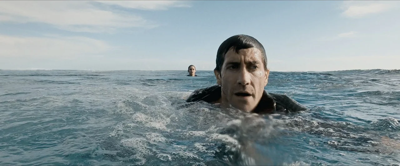 Road House (2024) movie still: medium — An open expanse of sea with gentle waves under a clear sky. A man swims in the foreground while a woman is…