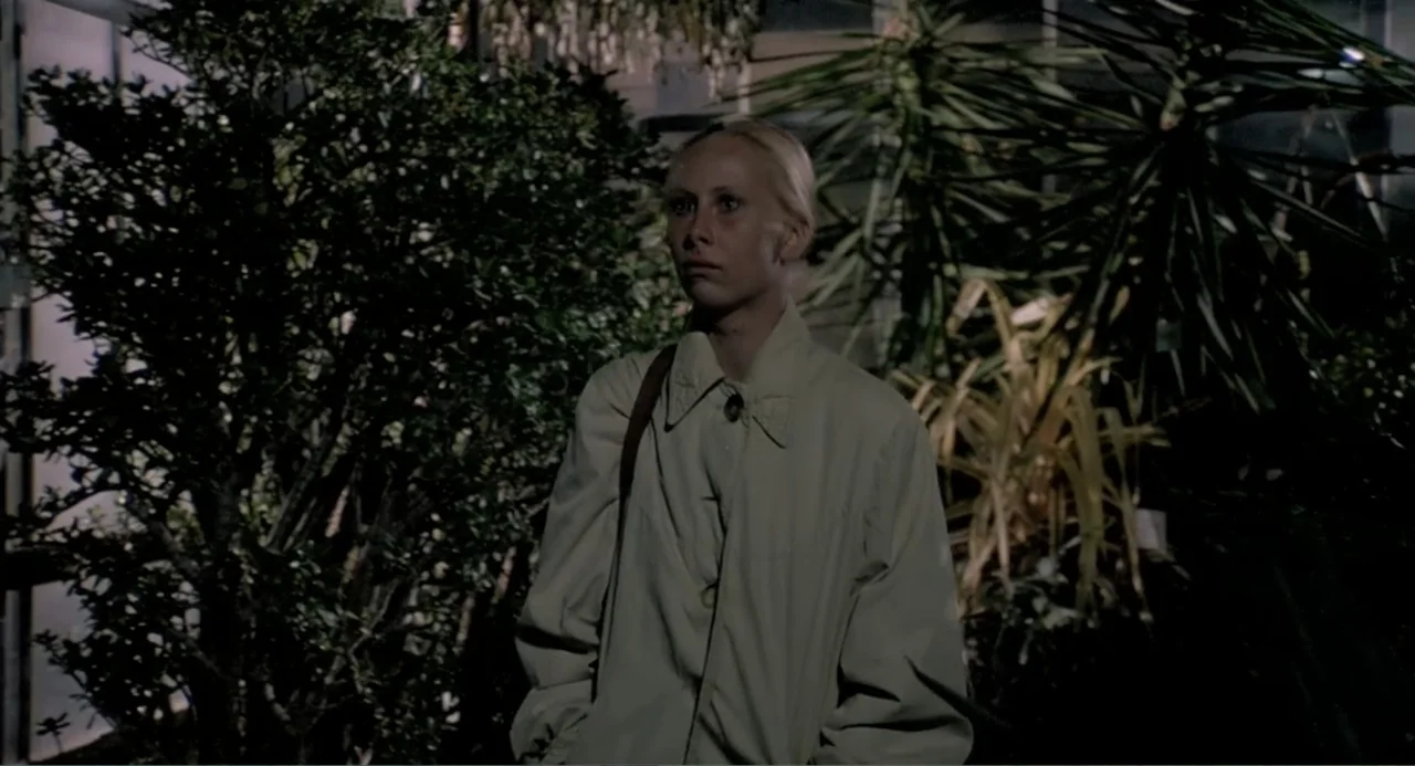 The Match Factory Girl (1990) movie still: medium — A dense greenhouse filled with various potted plants and foliage. A young woman in a trench coat…