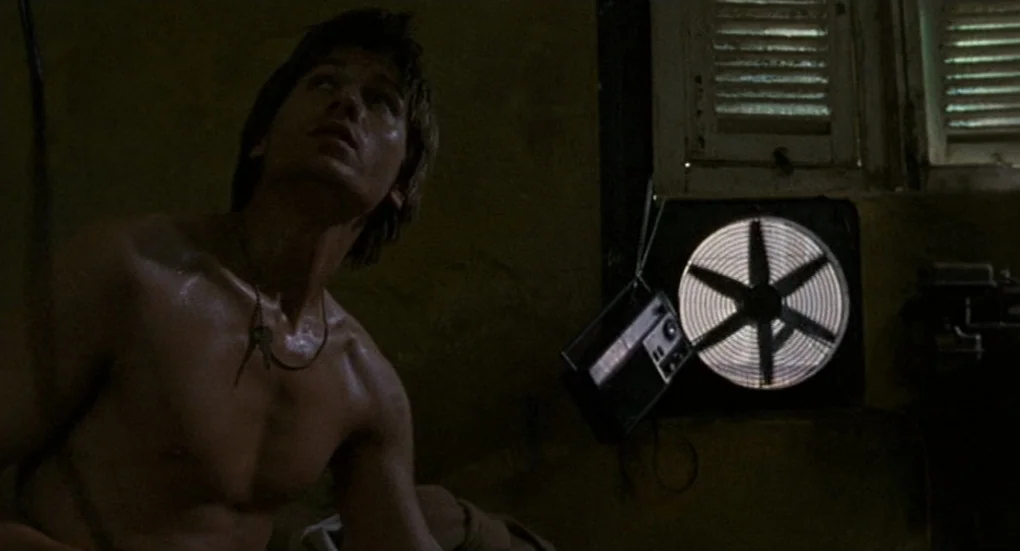 Furia (1999) movie still: medium — A dimly lit room with slatted window shutters and a small ventilation fan. A shirtless young man sits in the…