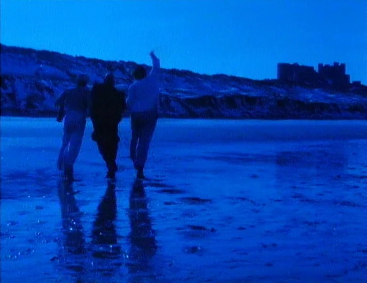 The Tempest (1979) movie still: wide — Three individuals walk across a wet, reflective beach under a dark blue night sky. A distant silhouette of a…