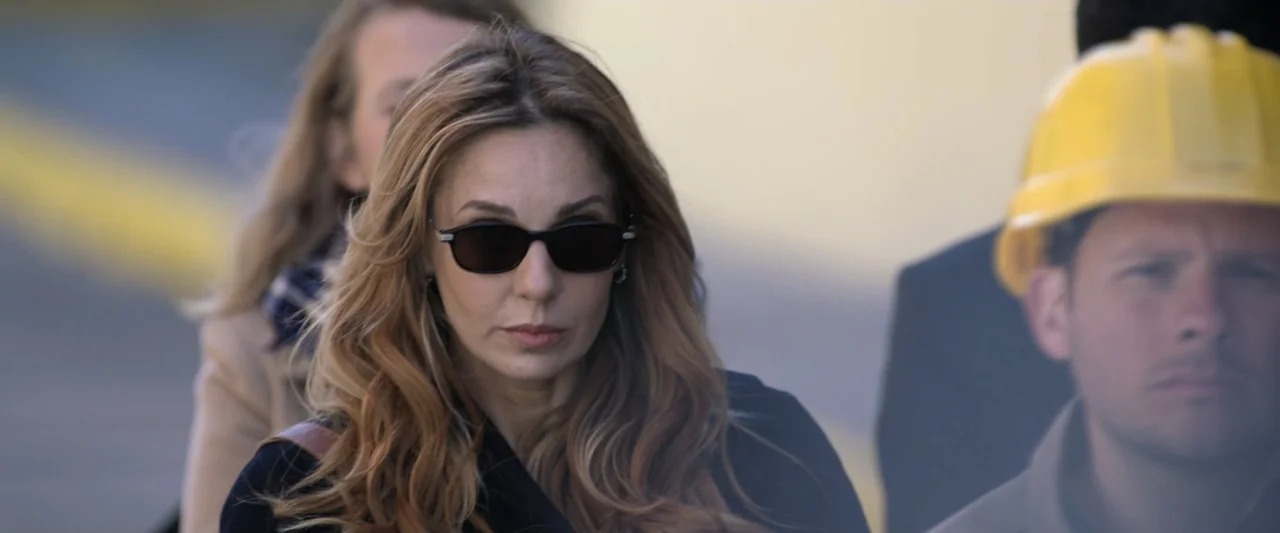 Doublespeak (2021) movie still: medium — An outdoor construction site with blurred background activity and bright daylight. A woman in sunglasses…