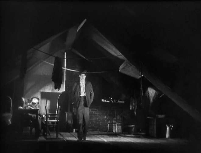 Pandora’s Box (1929) movie still: fullbody — A dark, cramped attic space features slanted walls and rough wooden floorboards. A young man stands in…