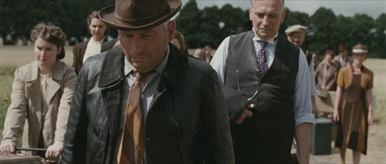 Suite Française (2015) movie still: medium — A group of people walk together along a path in an open countryside setting. A man in a leather hat…