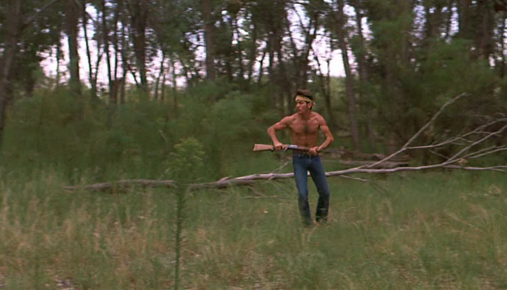 Badlands (1973) movie still: fullbody — An open patch of grass surrounded by dense pine trees and fallen branches. A shirtless young man holds a…