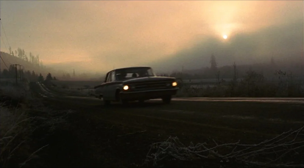 Five Easy Pieces (1970) movie still: wide — A vintage car travels along a desolate rural road surrounded by frosty vegetation at dawn. Thick,…
