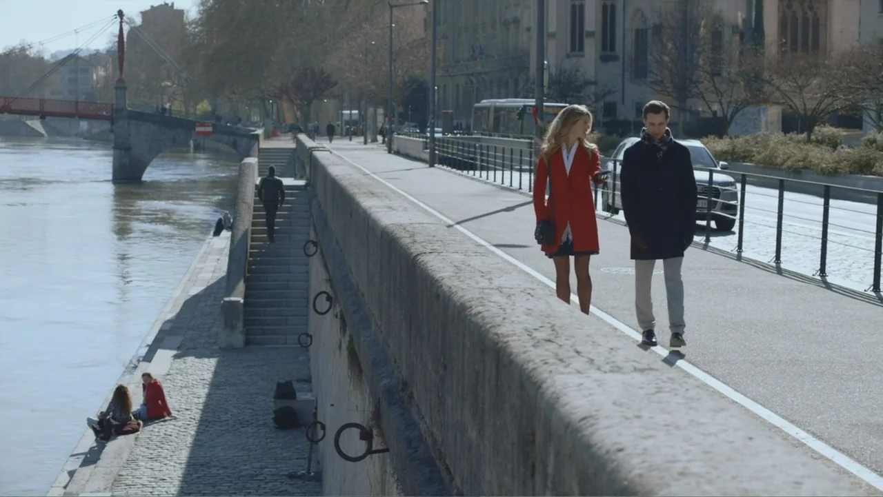 Driving Madeleine (2022) movie still: wide — A wide stone river embankment is populated by two people walking and another sitting. The pair walks…