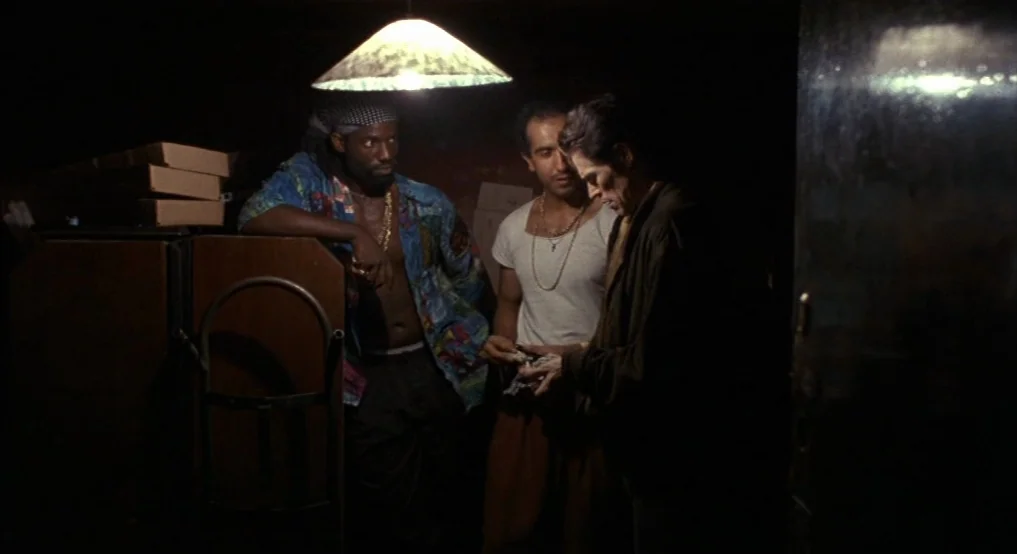 Light Sleeper (1992) movie still: medium — A dark, cluttered storeroom features cardboard boxes stacked on a metal dolly. Three men stand under a…