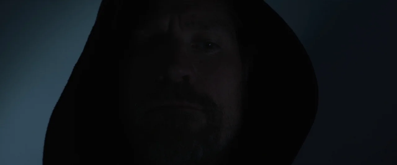 Small Crimes (2017) movie still: closeup — A man's face is partially obscured by shadows in an undefined, dark setting. He faces the camera directly…